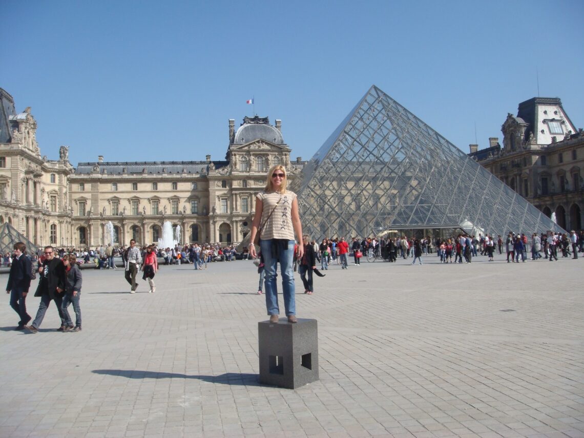 moça posando para foto em frente ao museu do louvre, com a piramide de vidro à direita e um dia de céu muito azul na primavera