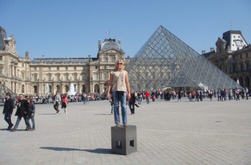 moça posando para foto em frente ao museu do louvre, com a piramide de vidro à direita e um dia de céu muito azul na primavera