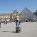 moça posando para foto em frente ao museu do louvre, com a piramide de vidro à direita e um dia de céu muito azul na primavera